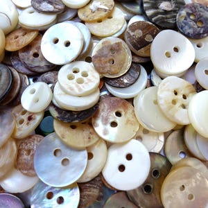 May include: A collection of assorted buttons in various shades of brown, white, and cream. The buttons are made of a smooth, shiny material, possibly mother-of-pearl or shell. Some buttons have two holes, while others have four holes.