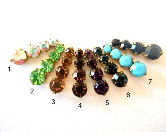 May include: A set of ten rhinestone embellishments in various colors, including iridescent, green, brown, purple, turquoise, and black. Each embellishment is a cluster of three rhinestones.