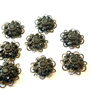 Op de afbeelding: Acht zwarte strassversieringen met een gouden metalen frame. Elke versiering is gevormd als een bloem met een centrale strasssteen omringd door kleinere strassstenen.