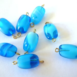 Puede incluir: Ocho cuentas de vidrio azul y blanco con lazos de metal dorado. Las cuentas son de forma ovalada y tienen un patrón de remolino.