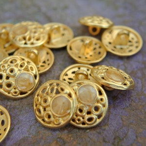 K&ouml;nnte beinhalten: Eine Sammlung goldfarbener Kn&ouml;pfe mit einem dekorativen, runden, durchbrochenen Design. Jeder Knopf hat eine kleine, runde, durchscheinende, hellfarbene Mitte.