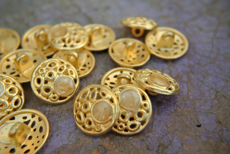 K&ouml;nnte beinhalten: Eine Sammlung goldfarbener Kn&ouml;pfe mit einem dekorativen, runden, durchbrochenen Design. Jeder Knopf hat eine kleine, runde, durchscheinende, hellfarbene Mitte.