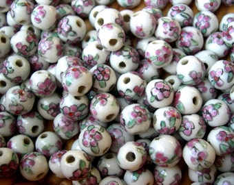 May include: A collection of white ceramic beads with pink and green floral designs. The beads are round and have a hole in the center for stringing.