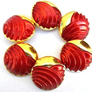 Op de afbeelding: Een set van vijf ronde knopen, elk met een gepolijste goudkleurige metallic afwerking aan de ene kant en een felrood, glanzend gedeelte met verhoogde, gebogen, schelpachtige ribbels aan de andere kant, gerangschikt in een cirkel op wit.