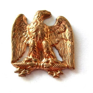 Puede incluir: Una figurita de águila de metal de color dorado con las alas extendidas. El águila está posada sobre una base con un diseño decorativo.