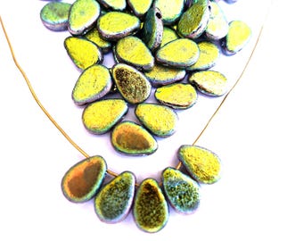 May include: A collection of iridescent, teardrop-shaped beads in shades of green, gold, and bronze. The beads are strung on a thin, gold-colored wire, with some beads scattered loosely. The beads have a metallic sheen and are arranged on a white background.