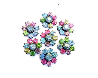 May include: A collection of six circular floral-shaped embellishments featuring a central round stone surrounded by a ring of smaller faceted crystals in various colors including light blue, pink, lime green, and yellow, set in dark metal bases.
