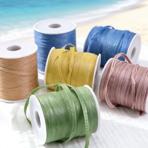 May include: A collection of colorful raffia ribbon spools in shades of blue, tan, yellow, pink, and green. Each spool is neatly wound and has a white core. The ribbons are arranged on a white surface with a blurred beach background.