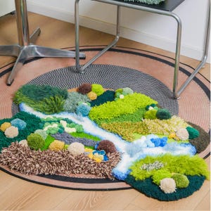K&ouml;nnte beinhalten: Ein handgefertigter Teppich mit einem Landschaftsdesign. Der Teppich zeigt einen Fluss aus Wei&szlig; und Blau, umgeben von getufteten Texturen in Gr&uuml;n, Braun und Gelb, die einer Miniaturlandschaft &auml;hneln. Der Teppich liegt auf einer runden, gemusterten Bodenbedeckung.