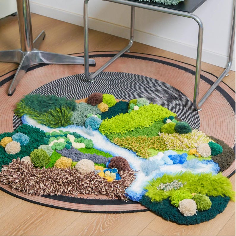 K&ouml;nnte beinhalten: Ein handgefertigter Teppich mit einem Landschaftsdesign. Der Teppich zeigt einen Fluss aus Wei&szlig; und Blau, umgeben von getufteten Texturen in Gr&uuml;n, Braun und Gelb, die einer Miniaturlandschaft &auml;hneln. Der Teppich liegt auf einer runden, gemusterten Bodenbedeckung.
