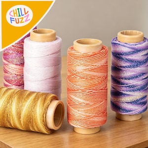 May include: A selection of colourful thread spools. The spools feature shades of pink, orange, purple, and gold. The thread is wound around wooden cores. The logo "CHILL FUZZ" is in the upper left corner.