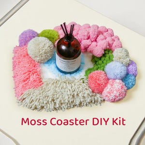 May include: A colorful DIY moss coaster kit with a brown aromatherapy bottle and reed diffuser sticks. The coaster features a variety of yarn pom-poms and a fluffy, textured base in shades of pink, green, blue, and purple. The text "Moss Coaster DIY Kit" is visible.