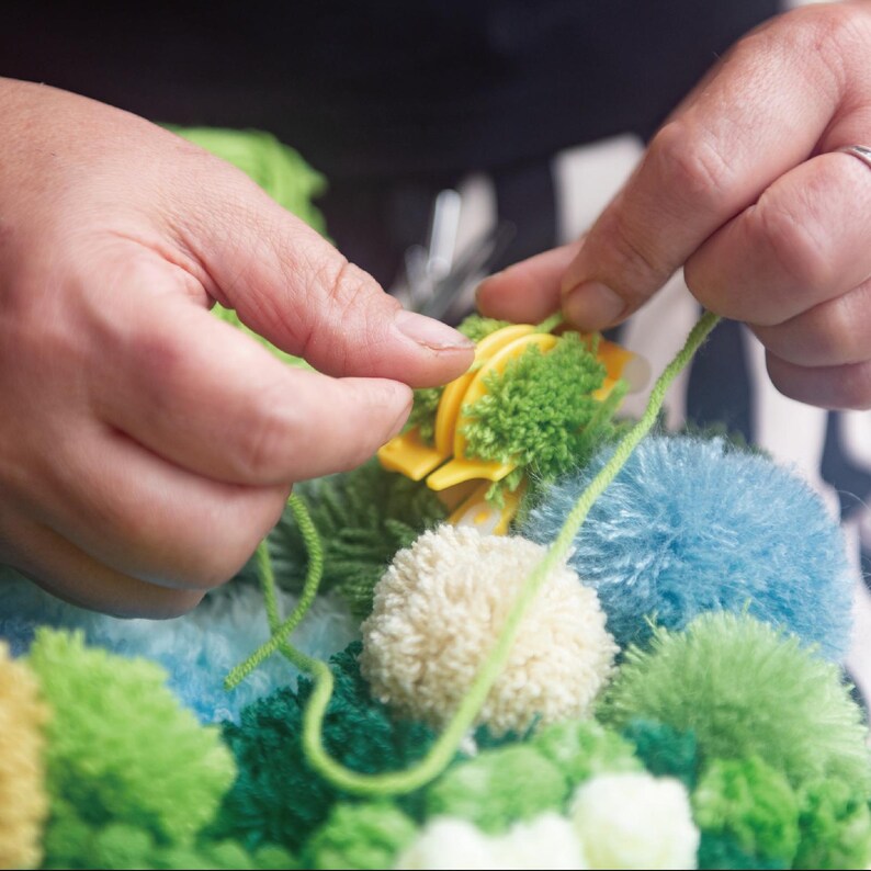 May include: Close-up of hands crafting a pom-pom. A yellow pom-pom maker is used with green yarn and fluffy pom-poms in various shades of green, blue, and cream. The craft project is in progress, with yarn being threaded through the maker.