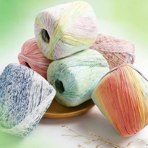 May include: A collection of colorful yarn skeins in various hues, including pink, blue, green, and yellow. The yarn is arranged in a pile, with some skeins resting on a small wooden plate. The background is a soft gradient of green and white.