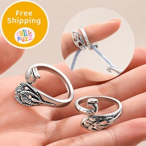 Könnte beinhalten: Zwei silberfarbene Metallringe mit Pfauenfeder-Design und einer offenen, verstellbaren Ringschiene. Ein Ring wird bei der Verwendung gezeigt, wie er einen hellblauen Garnfaden führt, während der andere separat abgebildet ist, um die Details zu zeigen.