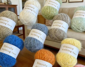 May include: A collection of colorful yarn skeins in various shades, including blue, green, yellow, and orange. Each skein has a white label with the text "Stuuuly Knitting Yarn". The yarn is arranged on a wooden surface.