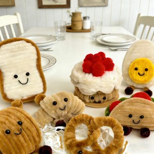 Puede incluir: Una colección de peluches de comida, que incluye una rebanada de pan tostado, una magdalena con pompones rojos, un huevo frito, un cruasán, un pretzel, un taco y una patata. Cada artículo tiene una cara sonriente.