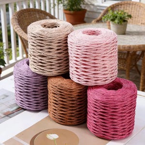 Può includere: Una collezione di rocchetti di spago di carta colorati nei toni del beige, rosa, viola, marrone e rosso. I rocchetti sono impilati, mostrando il loro aspetto strutturato e intrecciato. Lo spago è ideale per progetti di artigianato e decorativi.