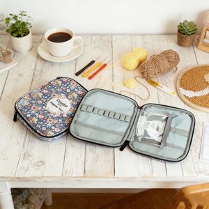 May include: An open, light blue craft organizer with multiple compartments, next to a closed floral print case. Crochet hooks, yarn, and a cup of coffee are also on the table. The text "Crocker Happier You" is visible on the closed case.