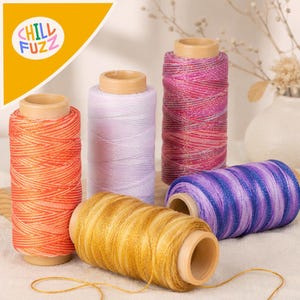 May include: Five spools of thread with metallic shimmer, including orange, light purple, pink and red variegated, gold, and purple and blue variegated colours. Each spool features a light brown cardboard core and wound thread.