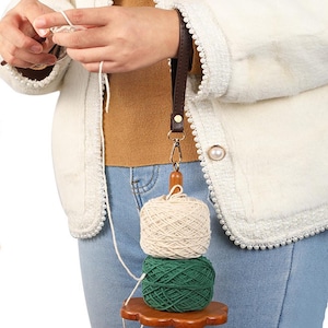 Könnte beinhalten: Ein Garnhalter mit einem braunen Lederriemen und einem Holzsockel. Der Halter hat zwei gestapelte Garnkugeln, eine cremefarbene und eine grüne. Der Halter ist mit einem goldfarbenen Verschluss an einer Handschlaufe befestigt.