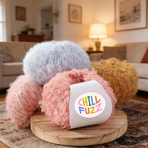 May include: Several balls of soft yarn in shades of pink, blue, and gold, displayed on a wooden board. A label on one ball says "CHILL FUZZ" in various colours. The yarn is suitable for knitting or crochet, perfect for crafting projects.