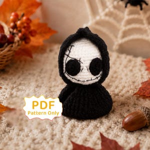 May include: A small crochet figure featuring a white skull-like head with large black circular eyes and stitched mouth details, wearing a black hooded cloak. A white label with the text PDF Pattern Only is positioned to the left of the figure.