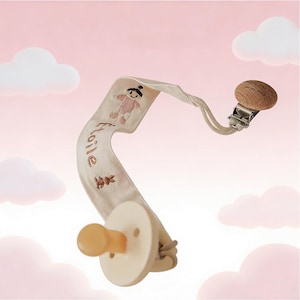 May include: A cream-colored baby pacifier with a matching fabric strap featuring embroidered teddy bear and bunny designs. The strap has the word "Etoile" embroidered on it. The pacifier has a wooden clip and a natural rubber nipple.