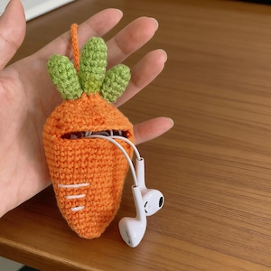 Könnte beinhalten: Eine handgefertigte orangefarbene Häkel-Karotten-Hülle mit grünen Blättern, die weiße Ohrhörer hält. Die Hülle dient zur Aufbewahrung und zum Schutz von Ohrhörern und bietet ein einzigartiges und verspieltes Accessoire.