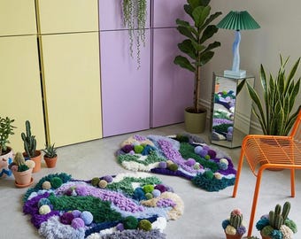 Puede incluir: Dos alfombras abstractas de forma orgánica, con una mezcla de texturas y colores. Las alfombras presentan tonos de morado, verde, azul y blanco, con hilos texturizados y detalles de pompones. Están sobre un suelo gris, con plantas en macetas y una silla naranja.
