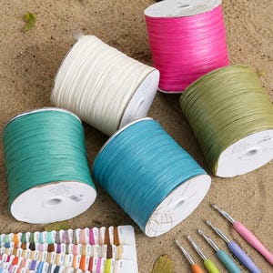 May include: Five spools of raffia ribbon in pink, white, olive green, teal, and turquoise, arranged with a color sample card and five crochet hooks with colored handles. The spools have white cardboard centers and a textured, fibrous appearance.