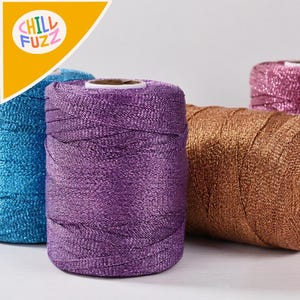 May include: Close-up of several spools of colorful yarn. The yarn is in shades of blue, purple, and gold. The spools are cylindrical and have a shiny, metallic appearance. The logo "CHILL FUZZ" is in the upper left corner.