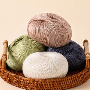 Peut inclure: Quatre pelotes de laine rondes de couleur rose poudré, vert sauge, crème et gris anthracite foncé sont disposées dans un panier rond en rotin tressé doté de deux anses en bois. Le fil présente une texture lisse et légèrement lustrée.
