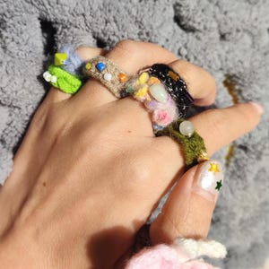 Puede incluir: Una mano que lleva varios anillos hechos a mano. Los anillos est&aacute;n hechos con hilo colorido, cuentas y otros adornos. Los anillos presentan varios dise&ntilde;os, incluyendo patrones florales y abstractos. Las u&ntilde;as est&aacute;n pintadas con una base blanca y dise&ntilde;os de estrellas.