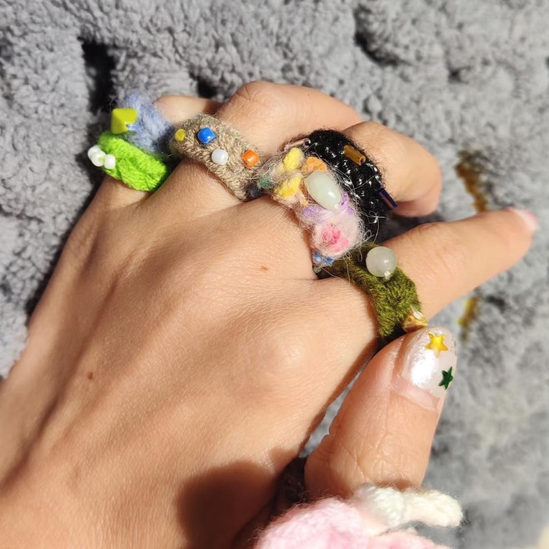 Puede incluir: Una mano que lleva varios anillos hechos a mano. Los anillos est&aacute;n hechos con hilo colorido, cuentas y otros adornos. Los anillos presentan varios dise&ntilde;os, incluyendo patrones florales y abstractos. Las u&ntilde;as est&aacute;n pintadas con una base blanca y dise&ntilde;os de estrellas.