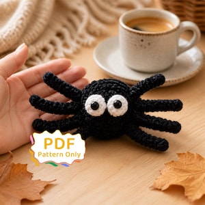 May include: A black crocheted spider toy with large white eyes and black pupils. The spider has eight legs and a round body. A white label with a scalloped edge features the text PDF Pattern Only in yellow and orange letters.
