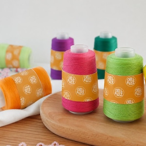 Puede incluir: Múltiples carretes de hilo en colores vibrantes que incluyen naranja, rosa, verde lima, morado y verde oscuro. Cada carrete presenta una banda de papel amarilla envuelta alrededor del centro con el texto Chill Fuzz impreso en blanco.