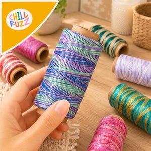 Puede incluir: Un carrete de hilo multicolor que presenta una mezcla de fibras metálicas azules, moradas y turquesas. El carrete se sostiene en primer plano, mientras que otros carretes de hilo de varios colores están dispuestos cerca sobre una superficie de madera.