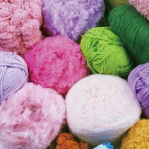 May include: A collection of colorful yarn balls and skeins in various shades of pink, purple, green, and white. The image showcases different textures, including fluffy, textured, and smooth yarn, perfect for knitting and crochet projects.