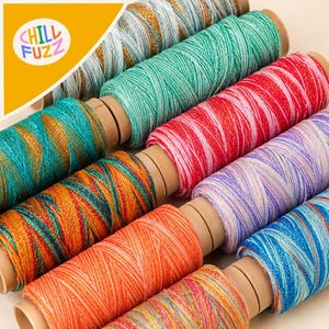 May include: A collection of multi-colored spools of thread arranged in rows. Each spool features vibrant, variegated yarn with metallic accents. The top left corner displays a yellow triangular graphic with the text CHILL FUZZ in colorful letters.
