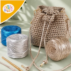 Puede incluir: Un bolso tipo cubo tejido a ganchillo en color dorado metálico con cierre de cordón, acompañado de tres ovillos de hilo brillante en azul, plata y oro, y dos ganchillos de metal. El logotipo CHILL FUZZ es visible en la esquina superior.