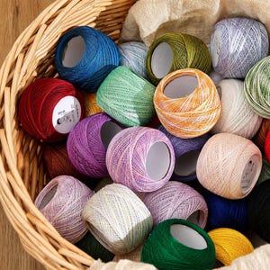 Op de afbeelding: Een verzameling veelkleurige haakgarenbollen in een gevlochten rieten mand. De garenbollen hebben diverse effen en gemêleerde kleuren, waaronder rood, blauw, groen, geel, paars, oranje en wit, elk voorzien van een centrale kern.