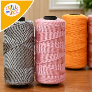 May include: Several spools of thread in shades of grey, pink, and orange. The thread is tightly wound on black bases. The image features the text "CHILL FUZZ" in a yellow and white design.