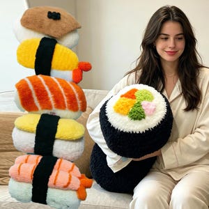 Op de afbeelding: Een verzameling pluche sushi-kussens in verschillende stijlen, waaronder nigiri en een makirrol. De nigiri-kussens hebben gele, oranje en roze elementen, terwijl het makirrol-kussen kleurrijke toppings heeft. De kussens zijn zacht en decoratief.