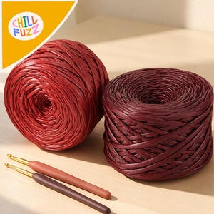 May include: Two round balls of ribbon yarn in shades of bright red and deep burgundy, accompanied by two crochet hooks with smooth handles in matching red and dark brown tones. The yarn has a flat, woven texture and a slight sheen.