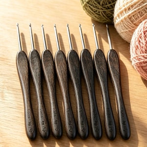 Puede incluir: Ocho ganchillos de ganchillo marrón oscuro con puntas de metal plateado. Los mangos de madera tienen un diseño tallado. Ovillos de lana verde, beige y rosa en el fondo, sugiriendo un tema artesanal.