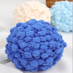 Puede incluir: Primer plano de una bola de hilo de pompón azul con una superficie texturizada. Otras bolas de hilo en crema y azul claro son visibles en el fondo. Este hilo es ideal para proyectos de manualidades.