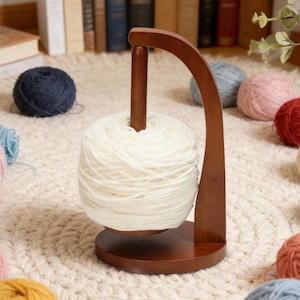 May include: A wooden yarn swift with a large ball of white yarn. The yarn swift is made of dark brown wood and has a curved arm that holds the yarn. Several balls of colorful yarn are scattered around the swift.