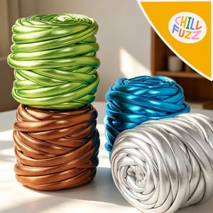 Può includere: Quattro rotoli di filato metallizzato verde, bronzo, blu e argento. Il filato è avvolto strettamente, creando un aspetto strutturato e stratificato. L'immagine include un logo che dice "CHILL FUZZ" in bianco, con un carattere arrotondato.