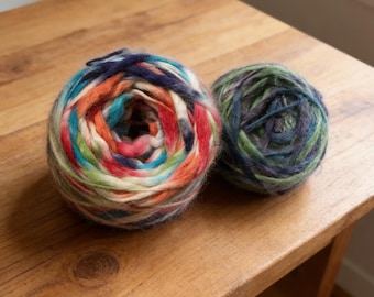 May include: Two balls of colourful yarn rest on a wooden surface. One ball displays a vibrant mix of colours including red, orange, blue, and green. The other ball is a mix of green, blue, and purple. The yarn appears soft and fluffy.